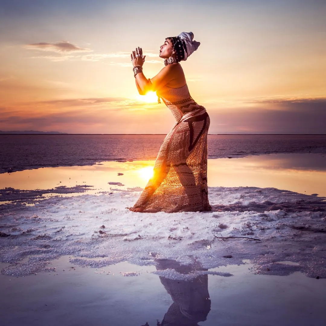 Donna su una spiaggia al tramonto, che unisce le mani in preghiera, simbolo di connessione spirituale, pace interiore e femminile sacro