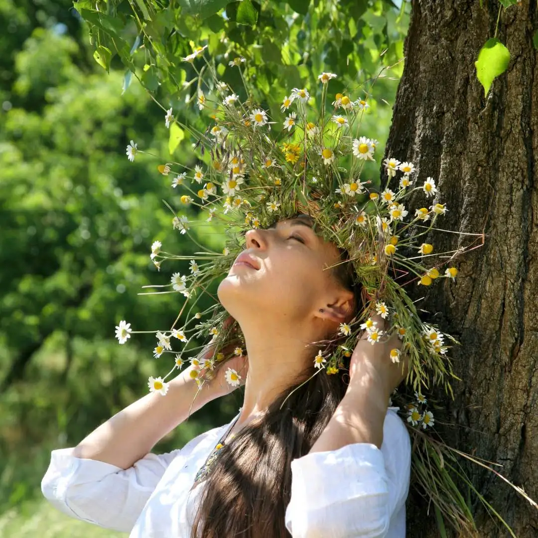 Frau genießt die milde Sonne, lehnt sich an einen Baum und trägt einen Blumenkranz, der die Verbindung zur Natur und innere Harmonie symbolisiert