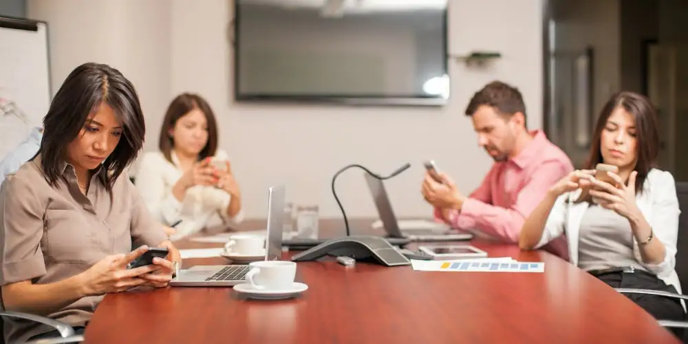 Personas en una sala de reuniones, distraídas con sus teléfonos celulares, ilustrando la dificultad de mantener la concentración frente a las distracciones modernas