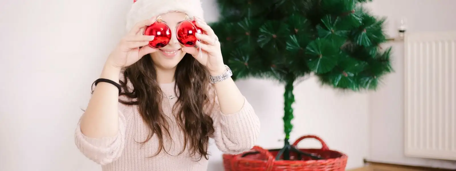 Woman in front of a Christmas tree, placing two baubles in front of her eyes, symbolizing the magic, wonder, and joy of celebrating the holidays
