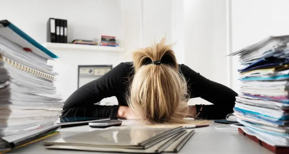 Mujer cansada en la oficina, con la cabeza sobre el escritorio con montones de documentos, simbolizando el agotamiento y la sobrecarga de trabajo